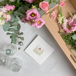 May include: A floral arrangement featuring pink roses, eucalyptus, and other greenery, presented in a cardboard box. A white square card with a watercolor illustration of a flower is also present, along with several glass vases.