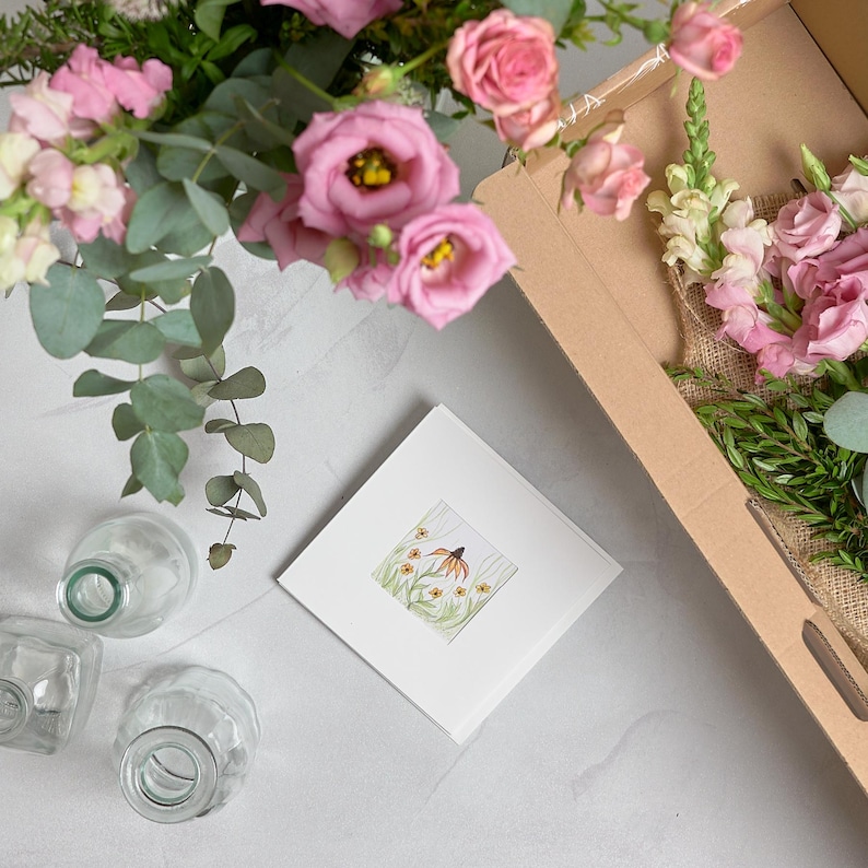 May include: A floral arrangement featuring pink roses, eucalyptus, and other greenery, presented in a cardboard box. A white square card with a watercolor illustration of a flower is also present, along with several glass vases.