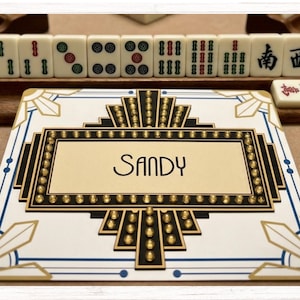 May include: A rectangular white card featuring a black and gold geometric frame with a sunburst design and small gold studs. The center of the card displays the name Sandy in a stylized black font. The card is positioned in front of mahjong tiles.