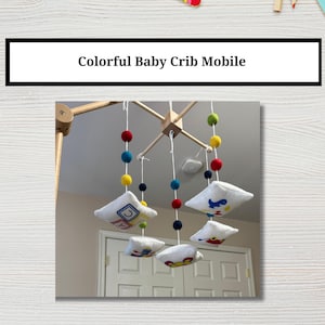 May include: Colorful baby crib mobile with five hanging fabric squares featuring different embroidered designs, including blocks, a dinosaur, and a car. The mobile is suspended from a wooden frame with colorful felt balls.
