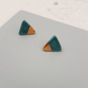 May include: A pair of teal and gold triangle stud earrings. Each earring features a teal triangle shape with a gold-coloured accent on one side. The earrings are presented on a light grey surface.