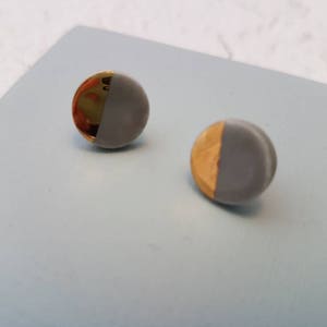 May include: Two round stud earrings. Each earring is half grey and half gold. The earrings are on a light blue surface.