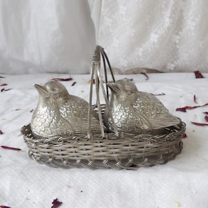 May include: A silver-toned salt and pepper set featuring two bird-shaped shakers nestled in a woven basket with a handle. The birds have detailed feather textures, adding a touch of elegance to the dining table.