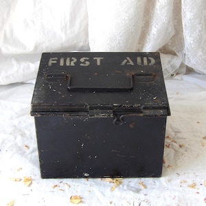 May include: A rectangular black metal first aid box featuring a hinged lid with a top handle and a front latch. The words FIRST AID are stencilled in a light, weathered font across the top surface of the lid. The metal shows signs of wear.