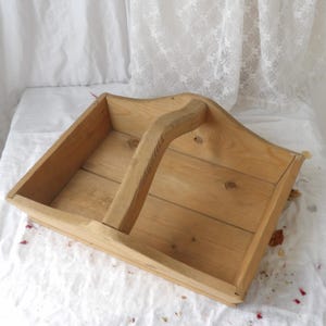 Pode incluir: Uma bolsa de madeira clara com uma alça. A bolsa tem dois compartimentos e é feita de madeira inacabada. A alça é curva e presa às laterais da bolsa. A bolsa está em uma superfície branca.