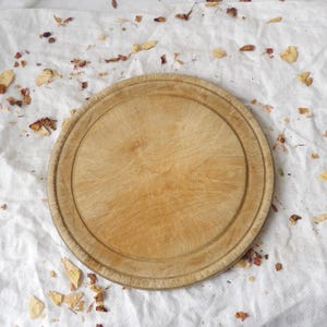 Op de afbeelding: Een ronde houten snijplank met een lichtbruine natuurlijke houtnerfafwerking. De plank is voorzien van een cirkelvormige verzonken groef nabij de buitenrand en vertoont zichtbare krassen op het oppervlak door gebruik.