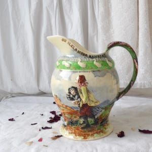 Könnte beinhalten: Keramikkrug mit cremefarbenem Ausguss und einem grünen Henkel mit rosa Akzenten. Der Korpus zeigt eine bemalte Szene mit Figuren und Text, darunter der Satz "ON ILKLA MOOR BAHT'AT". Der obere Rand hat einen grünen Rand mit weißen Entenillustrationen.