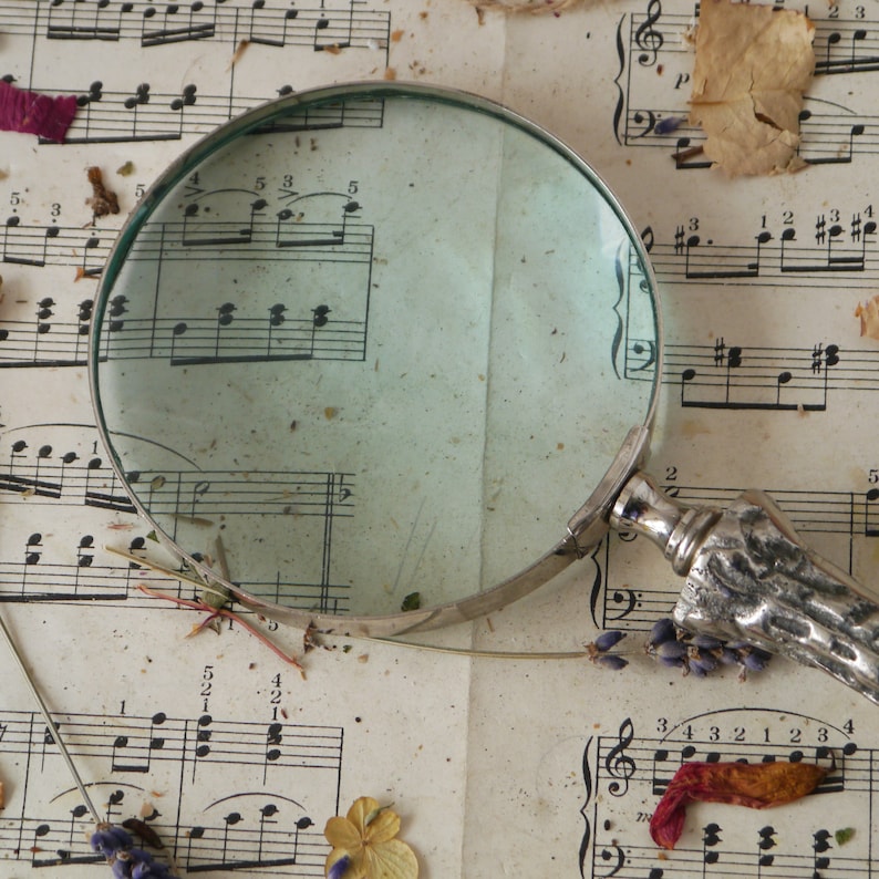 Peut inclure: Une loupe avec une poign&eacute;e argent&eacute;e repose sur une feuille de musique avec des notes et des chiffres musicaux. La loupe est positionn&eacute;e pour agrandir les notes de musique.