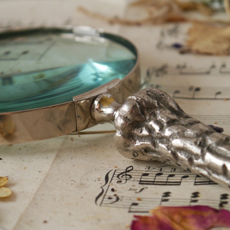 Peut inclure: Une loupe argent&eacute;e avec une lentille bleue et une poign&eacute;e textur&eacute;e d&eacute;taill&eacute;e. La loupe est pos&eacute;e sur une feuille de musique avec des notes musicales.
