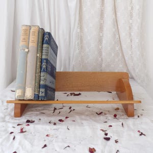 May include: A wooden bookshelf holds several vintage books with faded covers. The books are in shades of blue, green, and beige. The bookshelf is made of light-colored wood and has a simple, classic design.