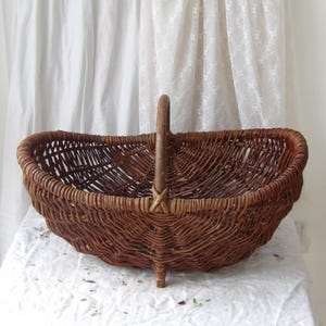 May include: A brown wicker basket with a handle. The basket has a rounded shape and is made of woven brown wicker. The handle is attached to the basket with a cross-shaped design. The basket has a sturdy base with small feet.