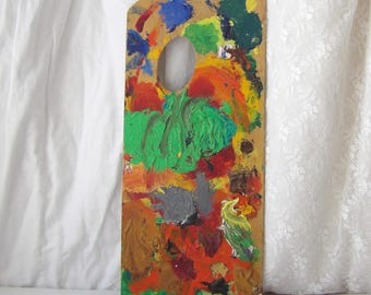 May include: A rectangular wooden artist palette featuring a thumb hole at the top. The surface is covered in thick, textured layers of dried oil paint in various colors including bright green, orange, red, yellow, blue, grey, and dark brown.
