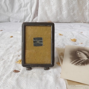 May include: A rectangular dark brown metal picture frame with rounded corners and two small feet at the base. The frame contains a textured mustard yellow insert with a central horizontal silver metal clip for holding a photograph in place.