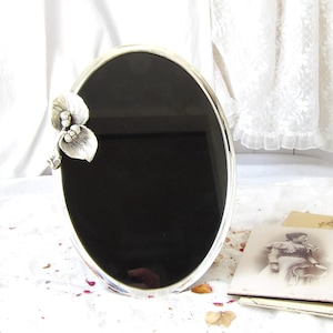 May include: An oval silver-coloured picture frame with a black interior. A decorative floral accent is on the upper left corner. The frame is on a white surface, with vintage photographs in the background.