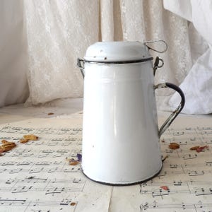 Op de afbeelding: Witte emaille kan met een deksel en een handvat. De kan heeft een versleten, vintage uitstraling.