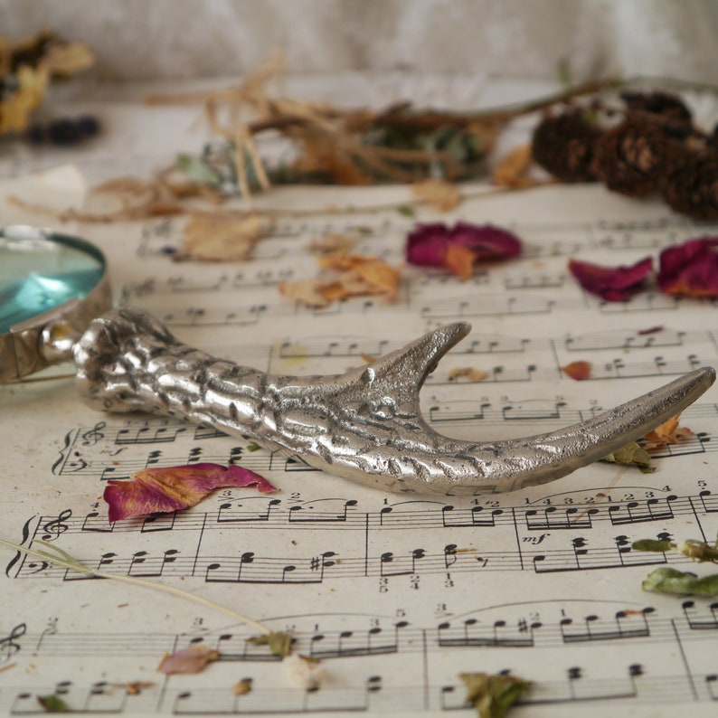 Peut inclure: Un ouvre-bouteille en argent en forme de bois de cerf avec un design d&eacute;taill&eacute;, reposant sur une feuille de musique avec des fleurs et des p&eacute;tales s&eacute;ch&eacute;s.