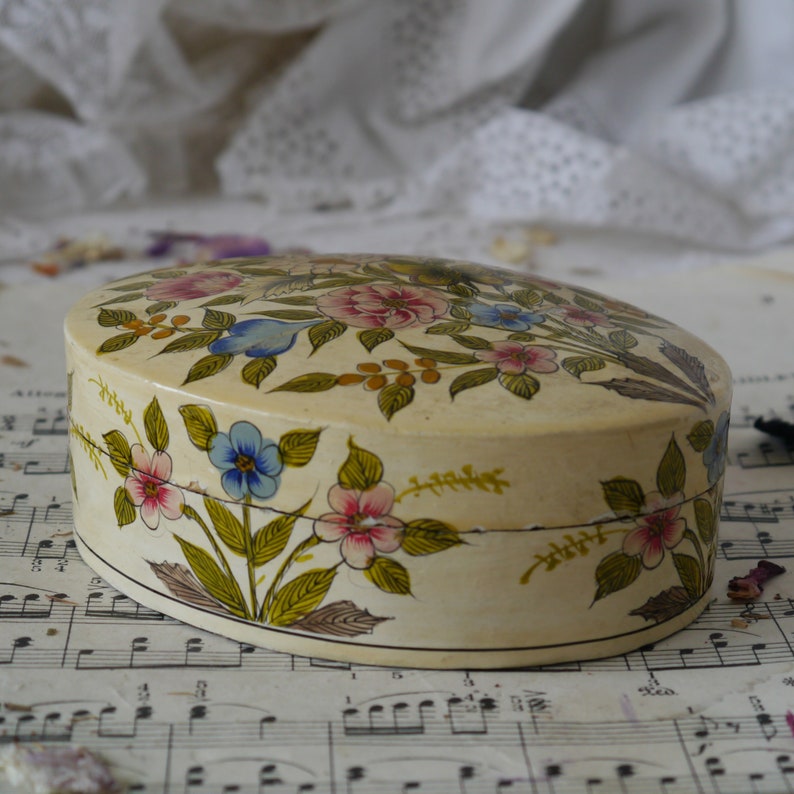 May include: An oval-shaped trinket box with a cream-colored base and a floral design painted in shades of pink, blue, yellow, and green. The box is sitting on a sheet of music.