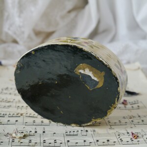 May include: A vintage, round, cream-colored trinket box with a floral design. The box has a black, lacquered bottom with a gold accent.