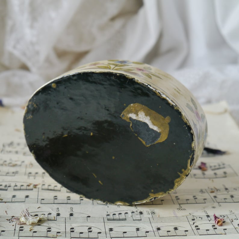 May include: A vintage, round, cream-colored trinket box with a floral design. The box has a black, lacquered bottom with a gold accent.