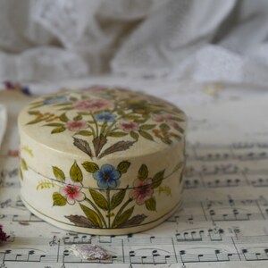 May include: A round, cream-colored trinket box with a floral design. The box is painted with pink, blue, and yellow flowers and green leaves.