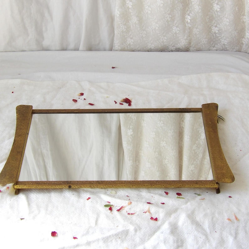 May include: A rectangular, vintage-style mirror with a gold-toned frame. The mirror has a simple design with curved supports on each end. The frame is made of wood and has a slightly distressed appearance. The mirror is resting on a white surface.
