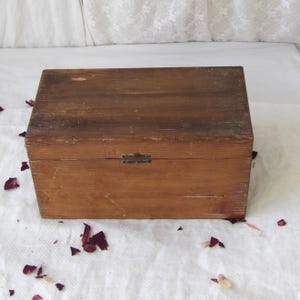 May include: A rectangular wooden box with a hinged lid and a metal clasp. The box is made of weathered, light brown wood with visible grain and some wear. The box is sitting on a white surface with scattered red rose petals.
