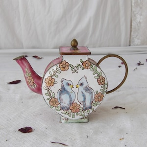 May include: A decorative teapot with a pink spout and handle, featuring a white body adorned with two blue birds and floral accents. The teapot has a gold-colored handle and a pink lid with a gold finial. A charming piece for display.