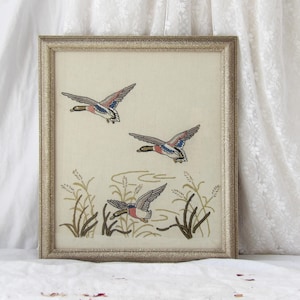 May include: Framed artwork featuring three embroidered ducks in flight above a pond with reeds. The ducks have gray, blue, and red wings. The artwork is in a gold-colored frame, creating a vintage aesthetic.