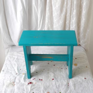 Puede incluir: Un pequeño taburete de madera pintado de color turquesa, con una parte superior rectangular y patas anguladas. El taburete tiene un acabado envejecido, que revela indicios de la veta de la madera y el color subyacente. Mide aproximadamente 30 cm de largo, 20 cm de ancho y 25 cm de alto.