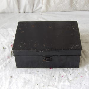 May include: A rectangular black metal box with a hinged lid and a clasp closure. The box has a slightly distressed appearance, with some areas showing wear and tear. The box is sitting on a white surface.