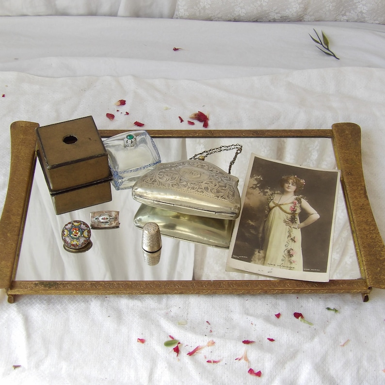 May include: An antique vanity mirror with a gold frame holds various vintage items. These include a small silver purse, a thimble, a small box, a ring box, a miniature mosaic, a small rectangular box, and a vintage photograph.