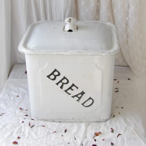 May include: White enamel bread bin, square with a matching lid. The word "BREAD" is printed in black on the front. The lid has a small, decorative handle. A vintage-style kitchen storage container.