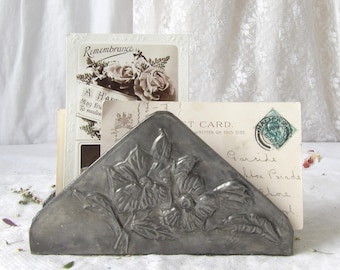 May include: A triangular, silver-colored metal card holder with a floral design. The holder is filled with vintage postcards, one with the text "Remembrance" and another with a stamp. The overall aesthetic is vintage and antique.