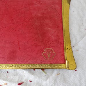 May include: A vintage red photo frame with a gold-colored metal border and accents. The frame has a textured red surface and a gold emblem in the lower right corner. The frame is rectangular with rounded corners.
