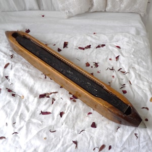 May include: A wooden weaving shuttle with a dark, rectangular interior. The shuttle is light brown with a pointed end and a metal tip. The item is resting on a white surface scattered with red petals.
