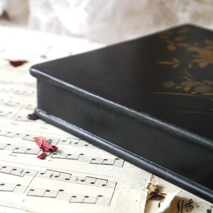 Pu&ograve; includere: Una scatola di legno nera con un disegno floreale dorato sulla parte superiore. La scatola &egrave; chiusa e si trova su un foglio di musica.