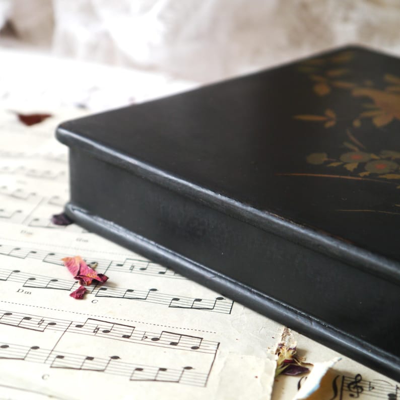 Pu&ograve; includere: Una scatola di legno nera con un disegno floreale dorato sulla parte superiore. La scatola &egrave; chiusa e si trova su un foglio di musica.