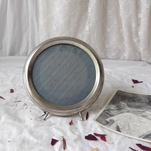Può includere: Una piccola cornice rotonda color argento con un retro in tessuto blu. La cornice ha un bordo perlato e tre piedini. Una fotografia in bianco e nero è accanto alla cornice. Petali di rosa sparsi sulla superficie bianca.