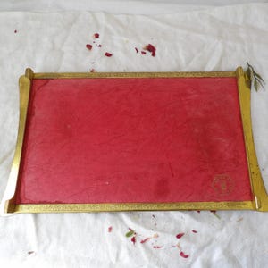 May include: A rectangular tray with a red interior and a gold-colored frame. The frame has decorative detailing and curved edges. A small gold emblem is in the lower right corner.