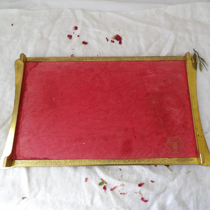 May include: A rectangular tray with a red interior and a gold-colored frame. The frame has decorative detailing and curved edges. A small gold emblem is in the lower right corner.