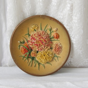 Può includere: Un piatto da parete rotondo, color beige, con un motivo floreale in rilievo. I fiori sono in tonalità di giallo, arancione e rosso, con foglie verdi. Il piatto ha un bordo marrone ed è probabilmente in ceramica.