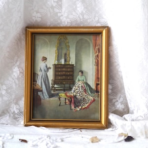 Op de afbeelding: Een ingelijste vintage print van een schilderij dat twee vrouwen in een kamer met een dressoir en een spiegel toont. Een vrouw staat en houdt een boek vast, terwijl de andere zit en een quilt naait.