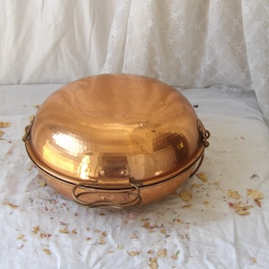 May include: A round, hammered copper cooking vessel with a domed lid. The pot features two brass-toned metal handles attached to the sides with rectangular plates and rivets, showcasing a polished metallic finish and a distinct textured surface.