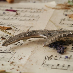 Peut inclure: Un ouvre-bouteille en m&eacute;tal argent&eacute; avec un design incurv&eacute; unique. L'ouvre-bouteille est pos&eacute; sur une feuille de musique avec des fleurs et des feuilles s&eacute;ch&eacute;es &eacute;parpill&eacute;es autour.