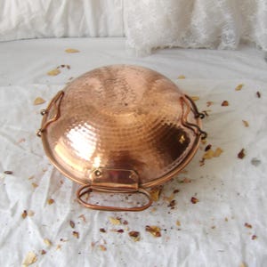 May include: A round, hammered copper bowl featuring a polished metallic finish and two curved side handles attached with rivets. The bowl has a wide, shallow basin shape with a smooth, reflective surface that catches light across its textured exterior.