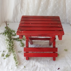 Puede incluir: Pequeña mesa plegable de madera roja, con tapa de listones. La mesa está pintada de rojo brillante y muestra signos de desgaste. Una ramita de flores blancas y hojas verdes descansa sobre la superficie blanca junto a la mesa.