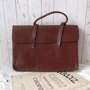 May include: A brown leather briefcase with a top handle and two shoulder straps. The briefcase is rectangular with a flap closure. A sheet of vintage sheet music is visible in the foreground.