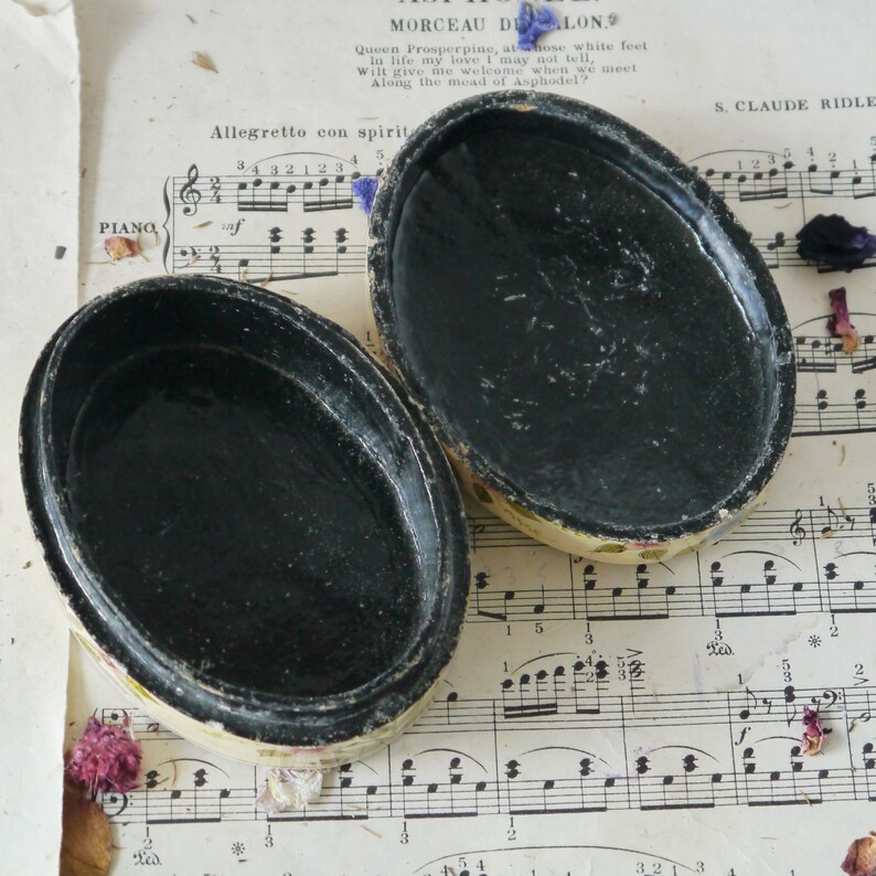 May include: A vintage oval-shaped trinket box with a black interior and a floral design on the exterior. The box is open and the lid is lying next to it. The box is sitting on a sheet of music.