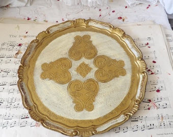May include: A round, ornate serving tray with a gold-painted rim and decorative gold accents on a cream-colored surface. The tray features a scalloped edge and is placed on sheet music.