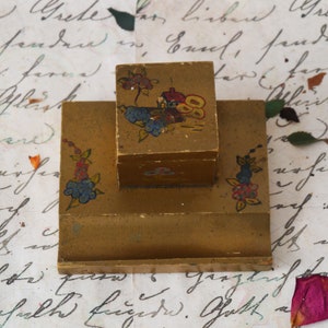 Może przedstawiać: Dwupiętrowy drewniany kałamarz z malowanym wzorem domu i kwiatów. Kałamarz jest pomalowany w odcieniach złota, brązu, czerwieni, błękitu i zieleni.